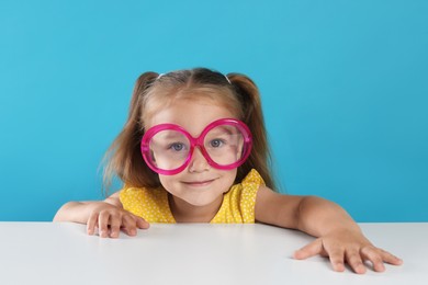 Photo of Curious little girl in funny glasses peeking over white table against light blue background