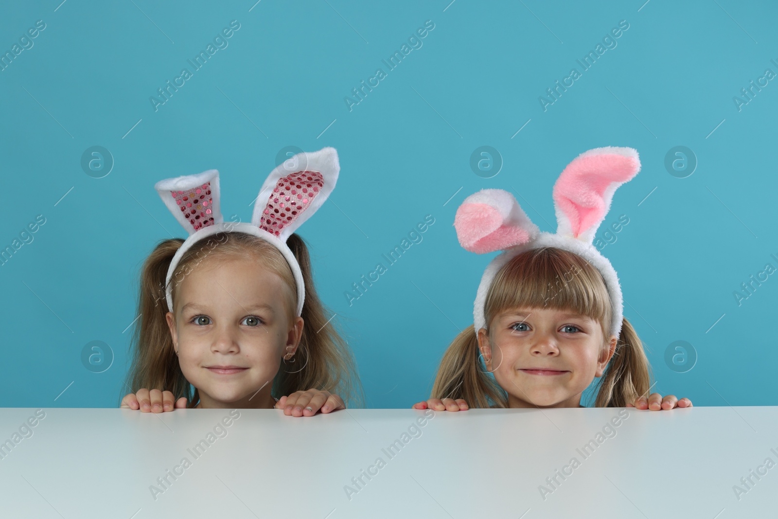 Photo of Curious little girls in bunny ears peeking over table against light blue background