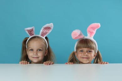 Photo of Curious little girls in bunny ears peeking over table against light blue background