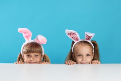Photo of Curious little girls in bunny ears peeking over table against light blue background