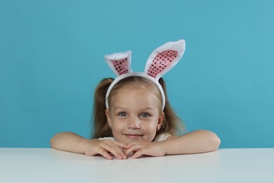 Photo of Curious little girl in bunny ears peeking over table against light blue background