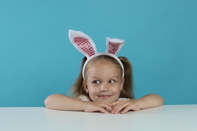 Photo of Curious little girl in bunny ears peeking over table against light blue background