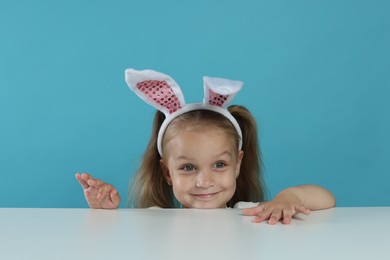 Photo of Curious little girl in bunny ears peeking over table against light blue background