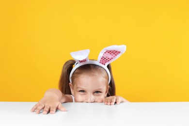 Photo of Curious little girl in bunny ears peeking over white table against orange background