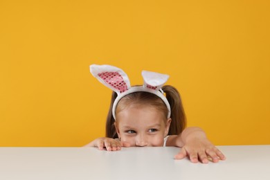 Photo of Curious little girl in bunny ears peeking over white table against orange background