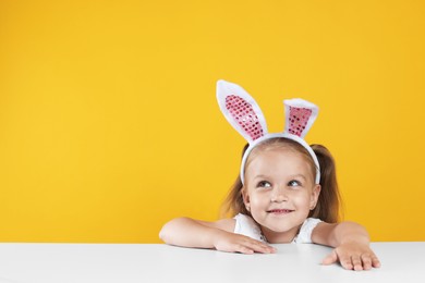 Photo of Curious little girl in bunny ears peeking over white table against orange background, space for text