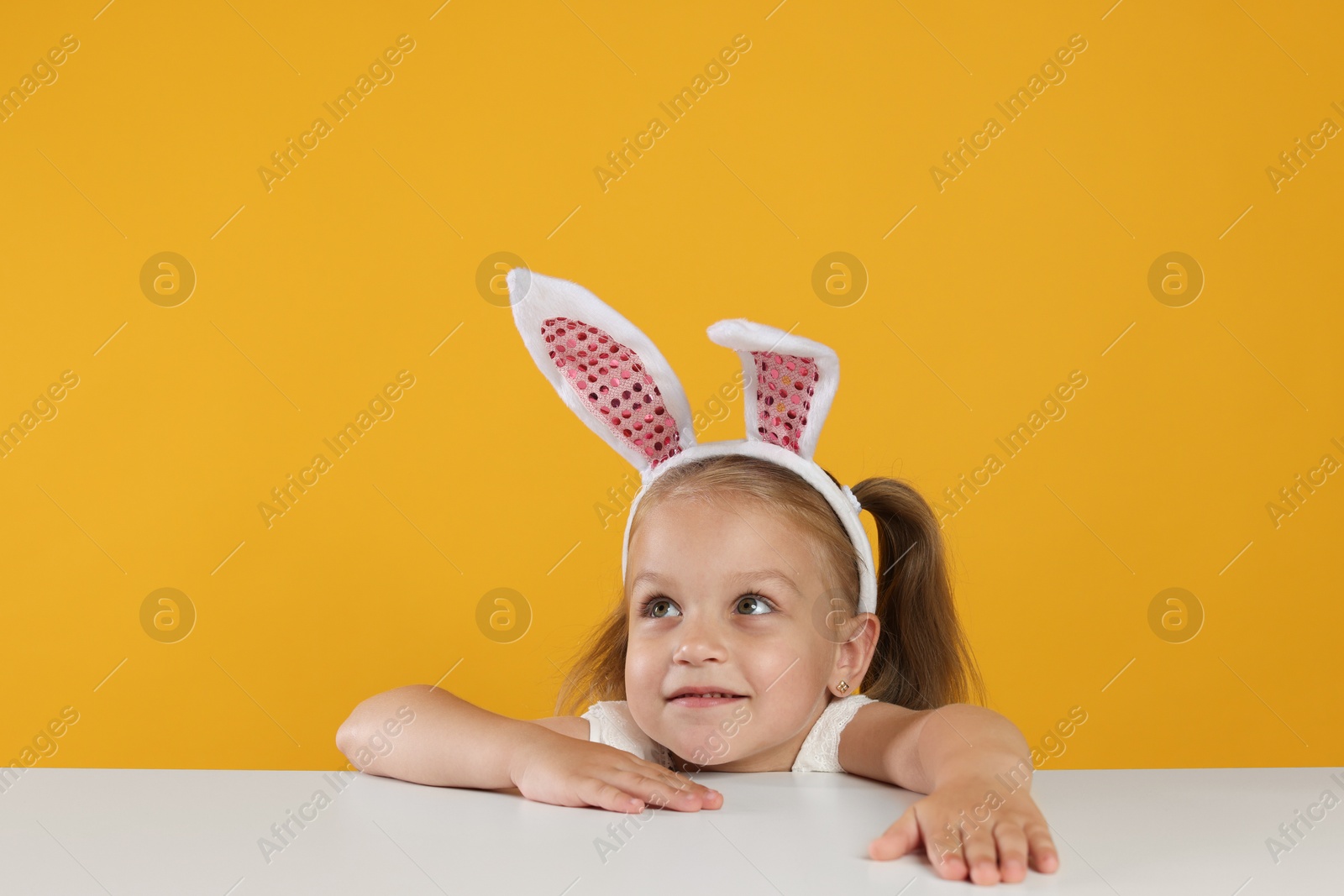 Photo of Curious little girl in bunny ears peeking over white table against orange background
