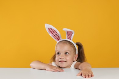 Photo of Curious little girl in bunny ears peeking over white table against orange background