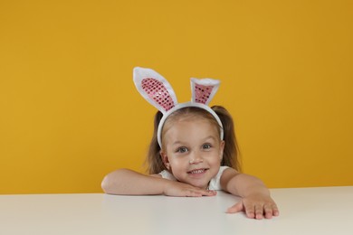 Photo of Curious little girl in bunny ears peeking over white table against orange background