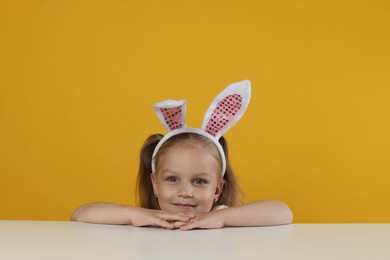 Photo of Curious little girl in bunny ears peeking over white table against orange background
