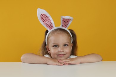 Photo of Curious little girl in bunny ears peeking over white table against orange background