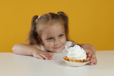 Photo of Cute little girl reaching for tasty dessert at white table against orange background, selective focus