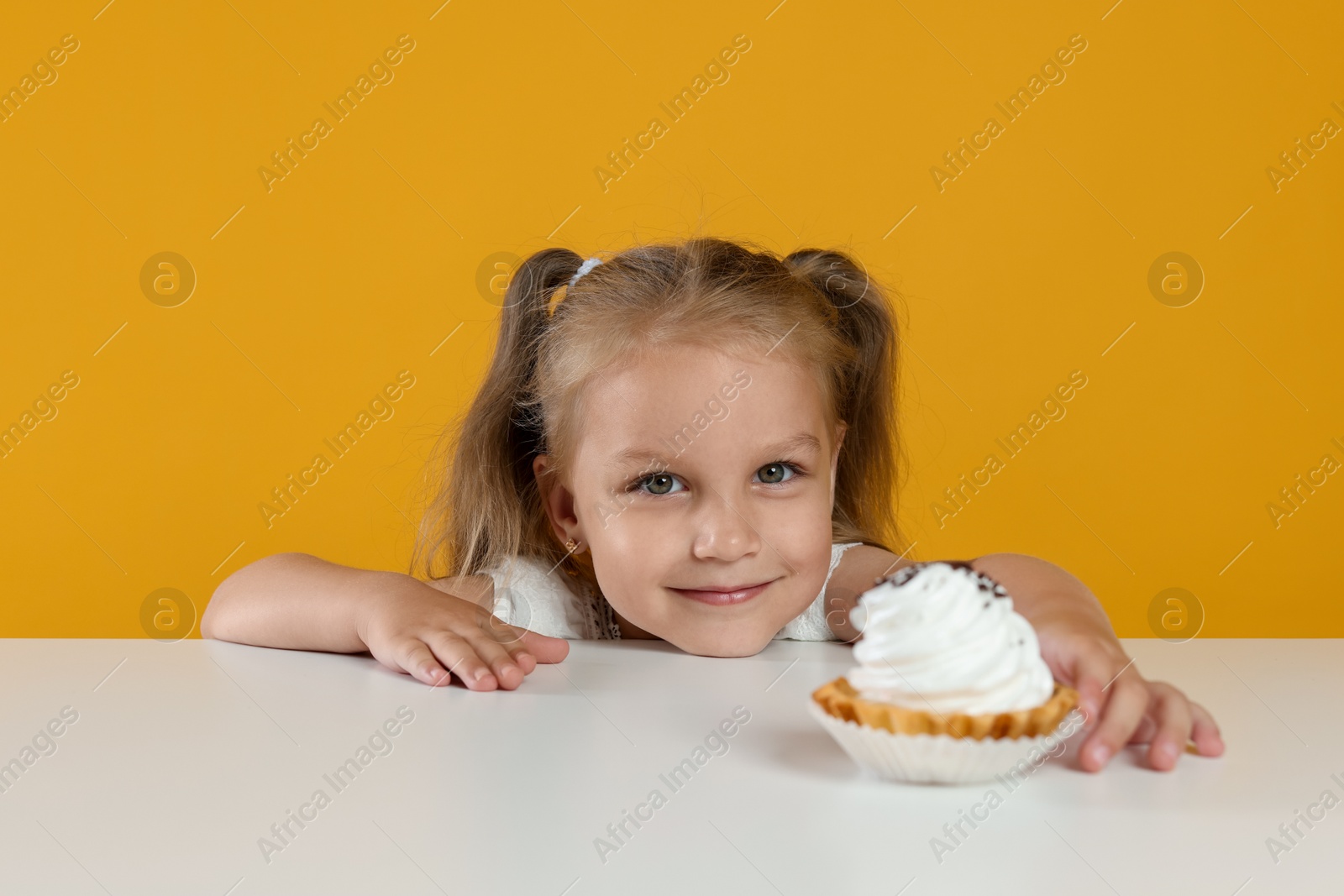 Cute little girl reaching for tasty dessert at white table against orange background Photo of Cute little girl reaching for tasty dessert at white table against orange background