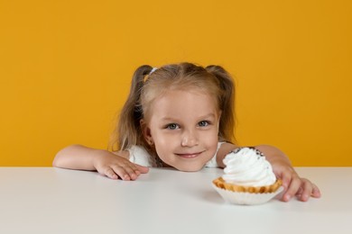 Photo of Cute little girl reaching for tasty dessert at white table against orange background