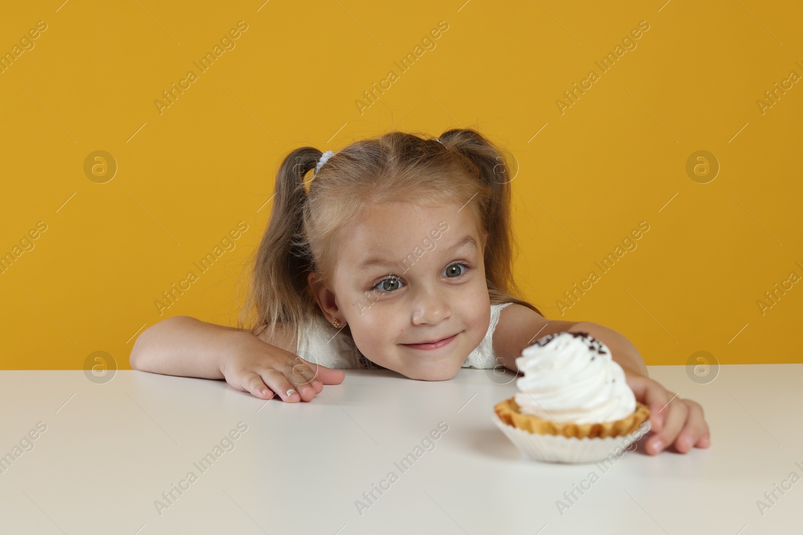 Photo of Cute little girl reaching for tasty dessert at white table against orange background