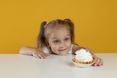 Photo of Cute little girl reaching for tasty dessert at white table against orange background