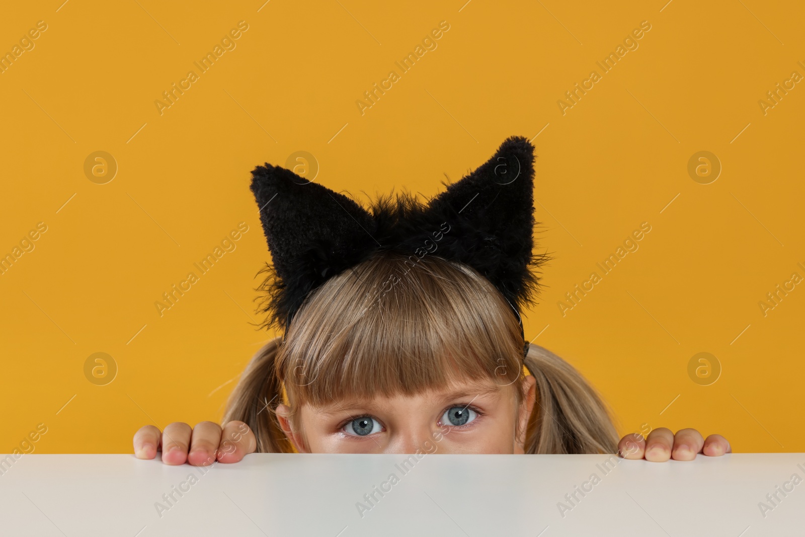 Photo of Curious little girl in cat ears peeking over white table against orange background