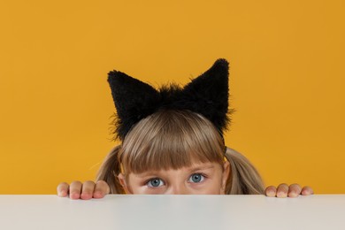 Photo of Curious little girl in cat ears peeking over white table against orange background