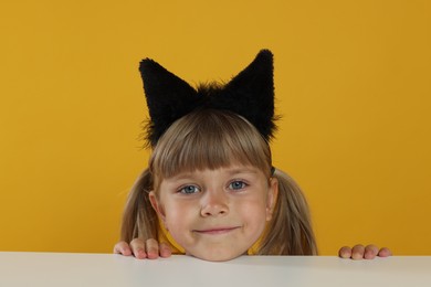 Photo of Curious little girl in cat ears peeking over white table against orange background