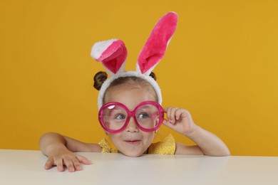 Photo of Curious little girl in bunny ears and glasses peeking over white table against orange background