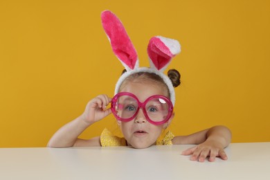 Photo of Curious little girl in bunny ears and glasses peeking over white table against orange background