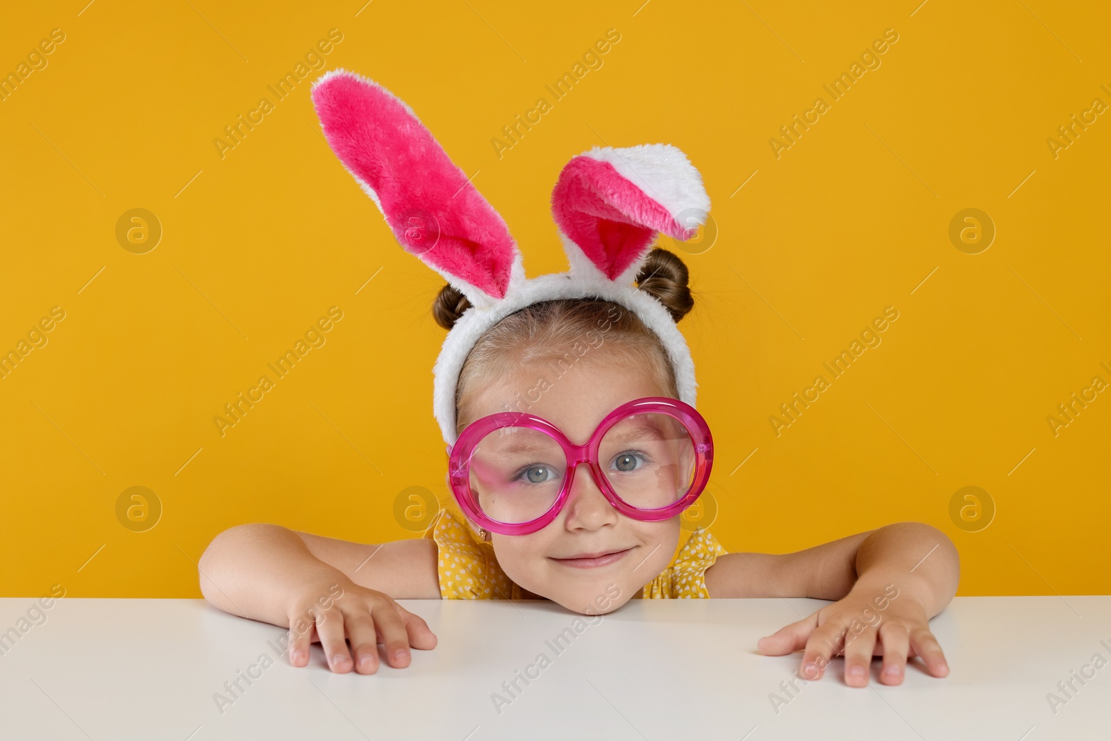 Photo of Curious little girl in bunny ears and glasses peeking over white table against orange background
