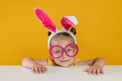 Photo of Curious little girl in bunny ears and glasses peeking over white table against orange background
