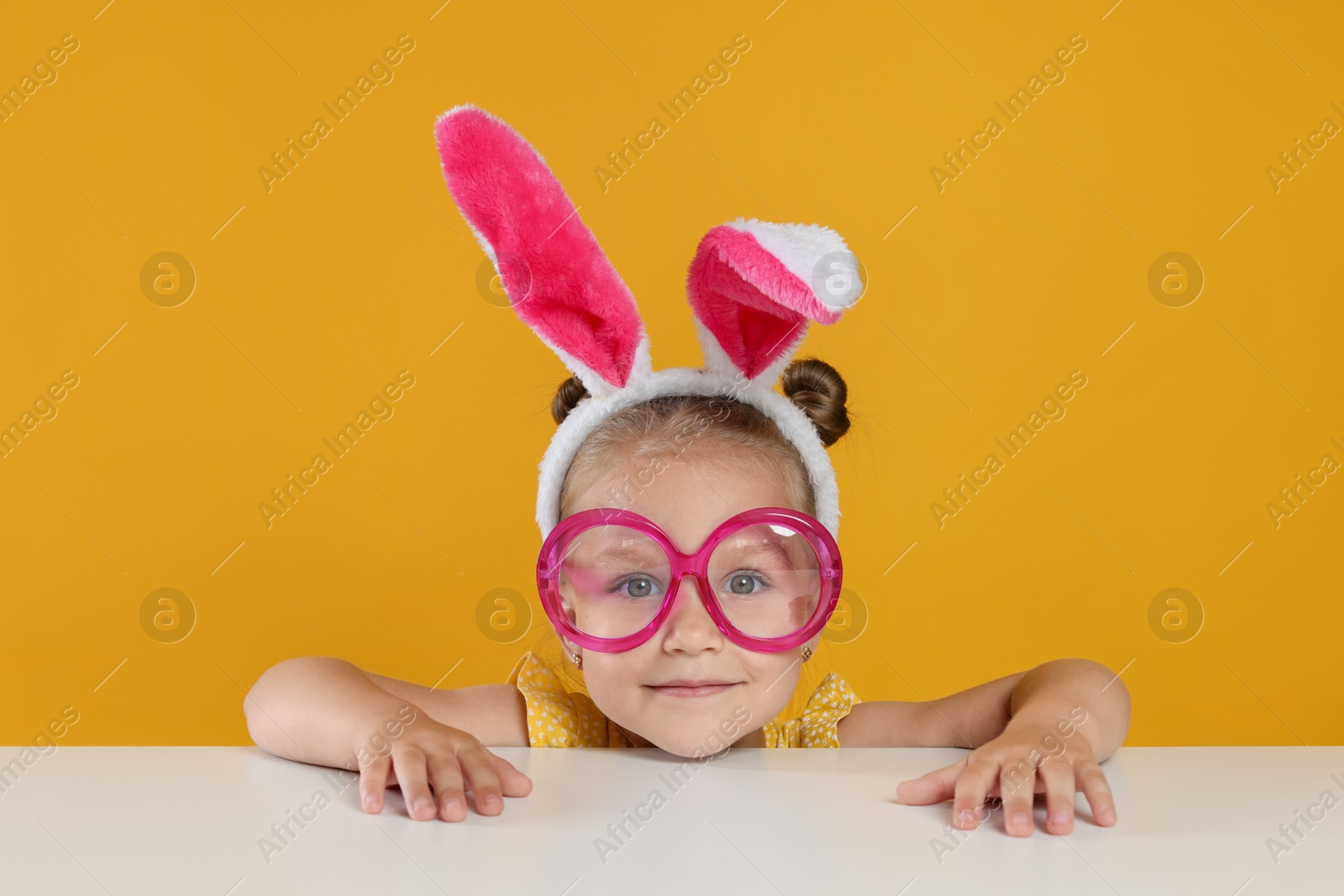 Photo of Curious little girl in bunny ears and glasses peeking over white table against orange background