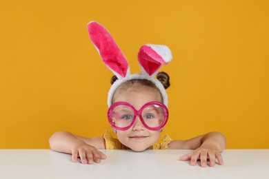 Photo of Curious little girl in bunny ears and glasses peeking over white table against orange background