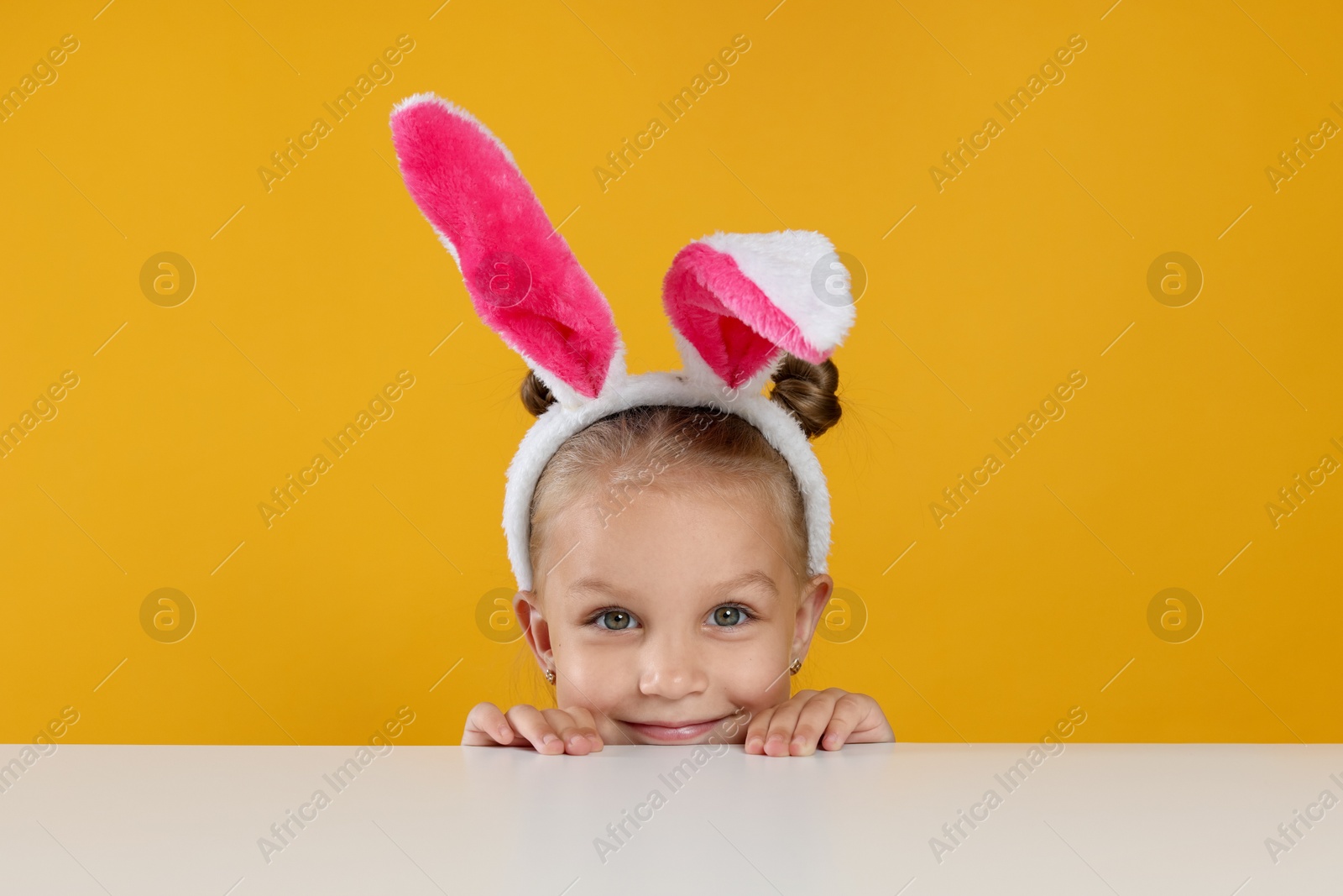 Curious little girl in bunny ears peeking over white table against orange background Photo of Curious little girl in bunny ears peeking over white table against orange background