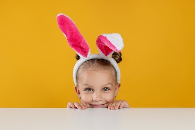 Photo of Curious little girl in bunny ears peeking over white table against orange background