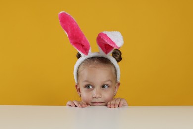 Photo of Curious little girl in bunny ears peeking over white table against orange background