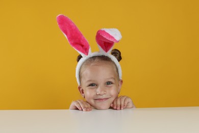 Photo of Curious little girl in bunny ears peeking over white table against orange background