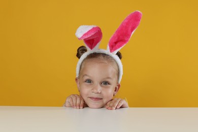 Photo of Curious little girl in bunny ears peeking over white table against orange background