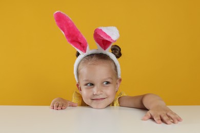 Photo of Curious little girl in bunny ears peeking over white table against orange background