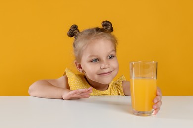 Photo of Cute little girl reaching for glass of juice at white table against orange background