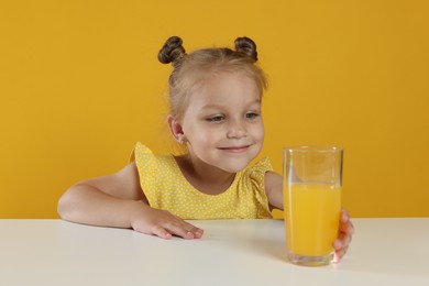 Photo of Cute little girl reaching for glass of juice at white table against orange background