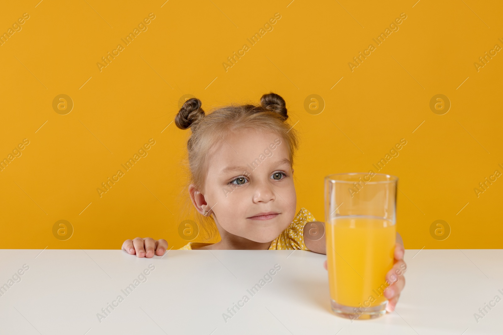 Cute little girl reaching for glass of juice at white table against orange background Photo of Cute little girl reaching for glass of juice at white table against orange background