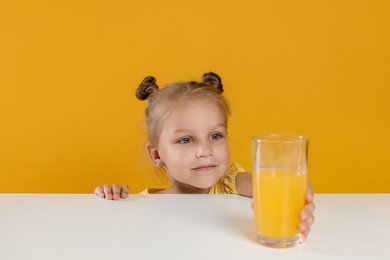 Photo of Cute little girl reaching for glass of juice at white table against orange background