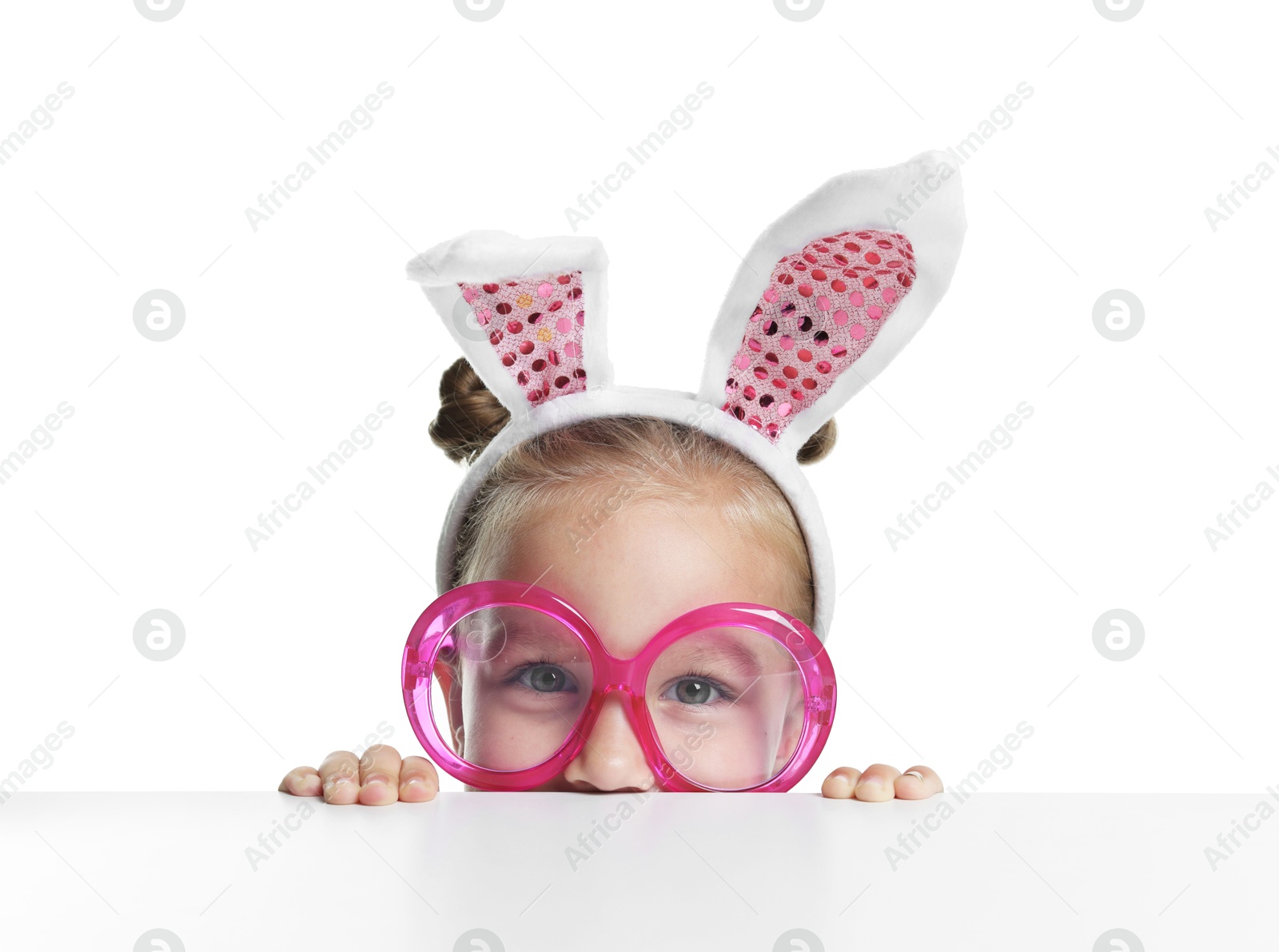 Photo of Curious little girl in bunny ears and glasses peeking over table against white background
