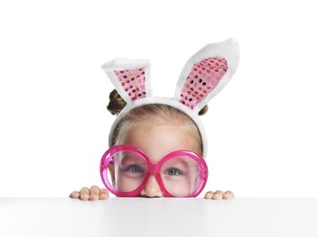 Photo of Curious little girl in bunny ears and glasses peeking over table against white background