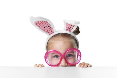 Photo of Curious little girl in bunny ears and glasses peeking over table against white background
