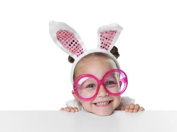 Photo of Curious little girl in bunny ears and glasses peeking over table against white background