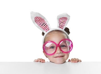 Photo of Curious little girl in bunny ears and glasses peeking over table against white background
