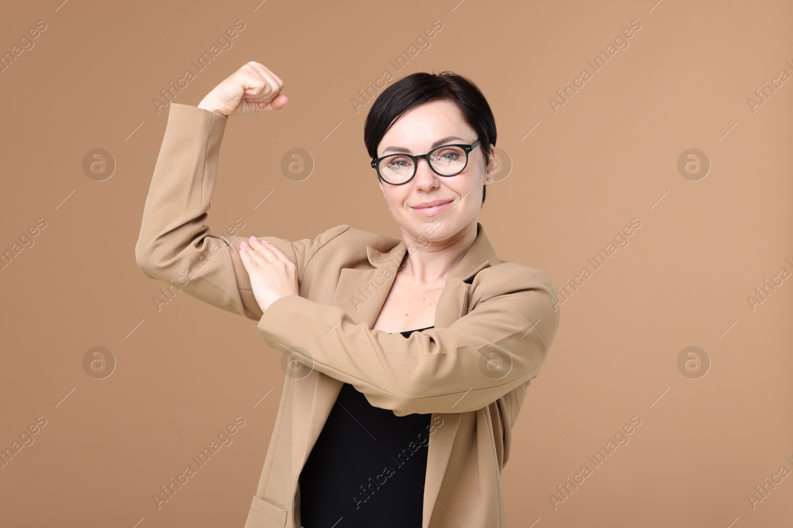 Photo of Strength. Beautiful businesswoman in glasses and suit showing her biceps on beige background