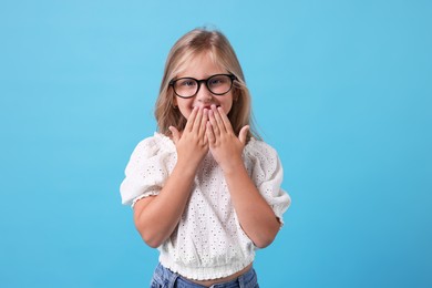 Photo of Cute little girl in glasses covering mouth and laughing on light blue background