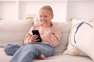 Photo of Cute little girl with smartphone covering mouth and laughing on sofa at home
