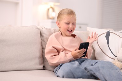 Photo of Cute little girl with smartphone laughing on sofa at home