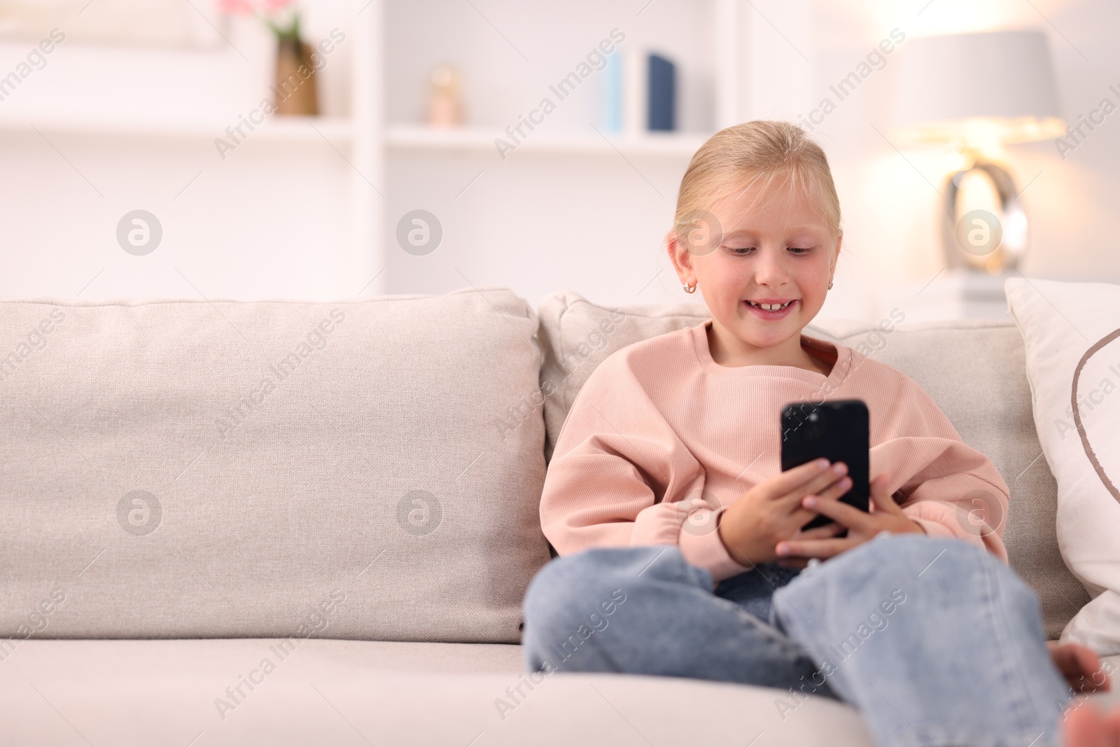 Photo of Cute little girl with smartphone laughing on sofa at home. Space for text