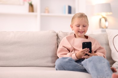 Photo of Cute little girl with smartphone laughing on sofa at home. Space for text